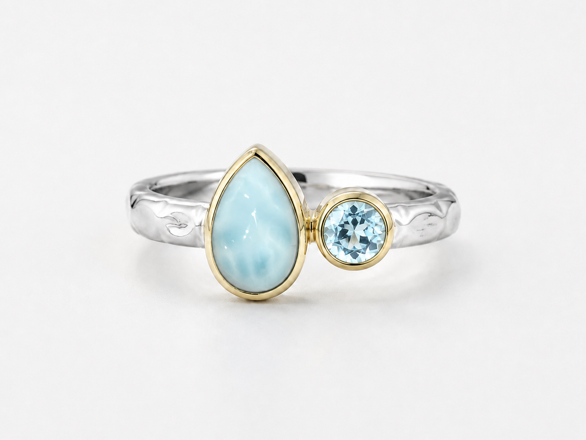 A silver ring with a teardrop-shaped light blue larimar stone and a small round light blue gemstone in gold bezels, set on a white background.