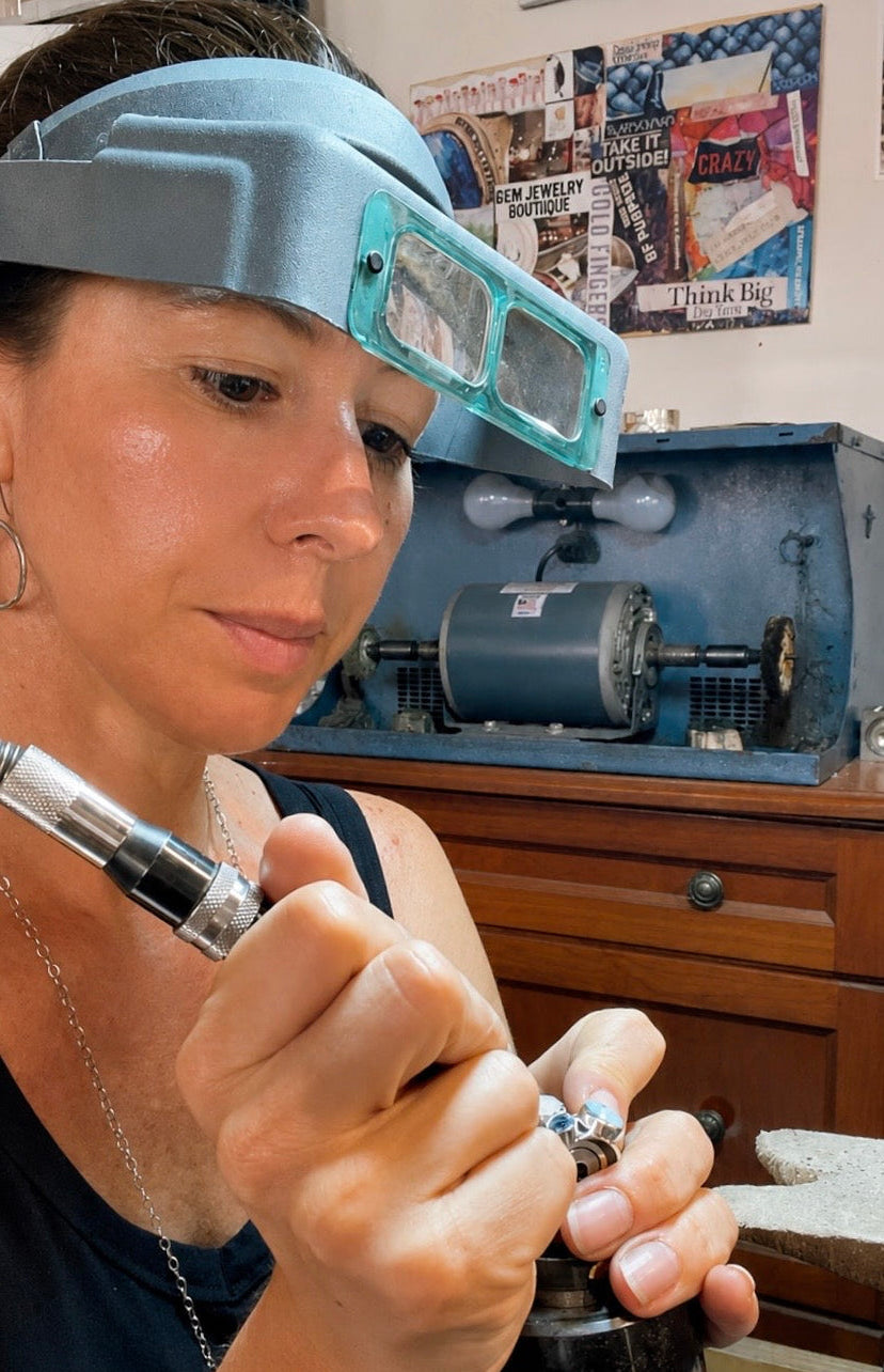 A close-up lifestyle shot of a jeweler wearing a magnifying headlamp and holding a silver ring under a jeweler's loupe, with a workshop background containing tools and a poster.